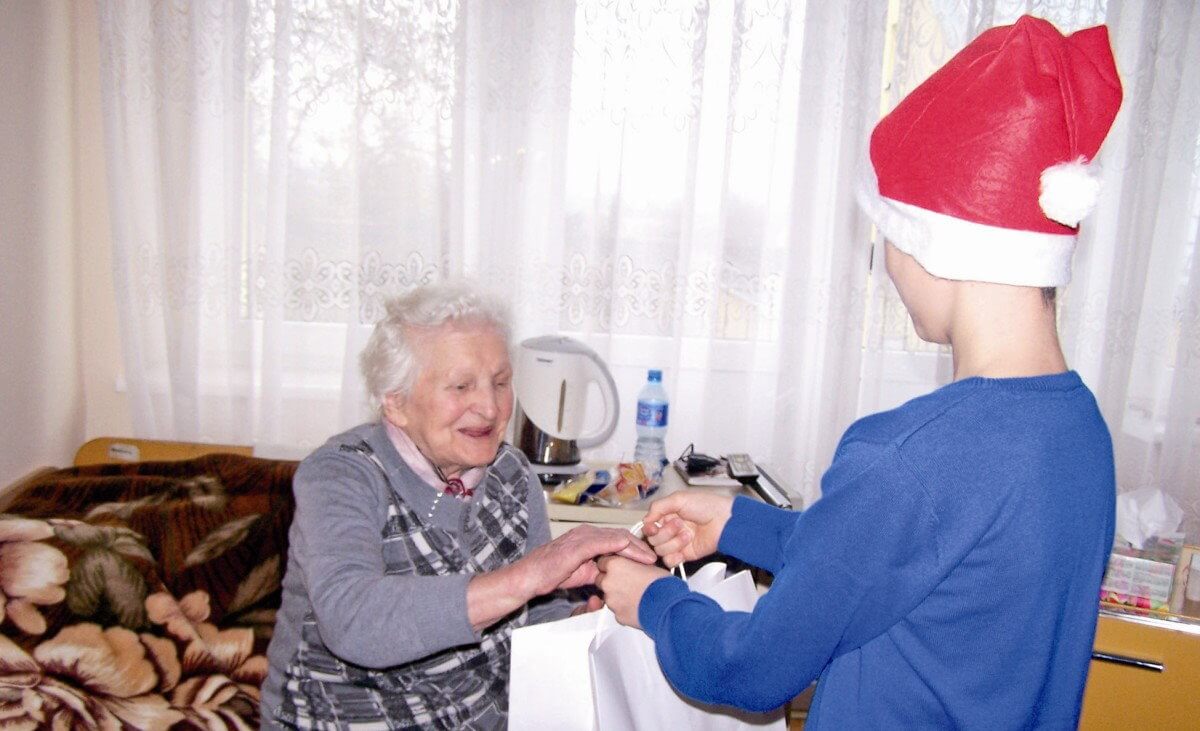 Dziecko z czapką Świętego Mikołaja na głowie wręcza paczkę seniorce