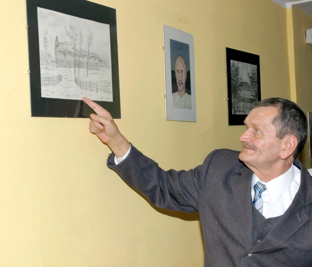 Leszek Czajka pokazuje palcem czarno-biały rysunek wiszący na ścianie