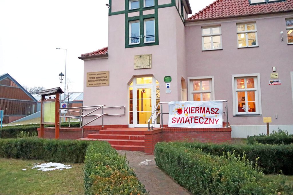 Siedziba Warsztatu Terapii Zajęciowej; na poręczy jest zawieszona biała plansza z czerwonym napisem „Kiermasz świąteczny”