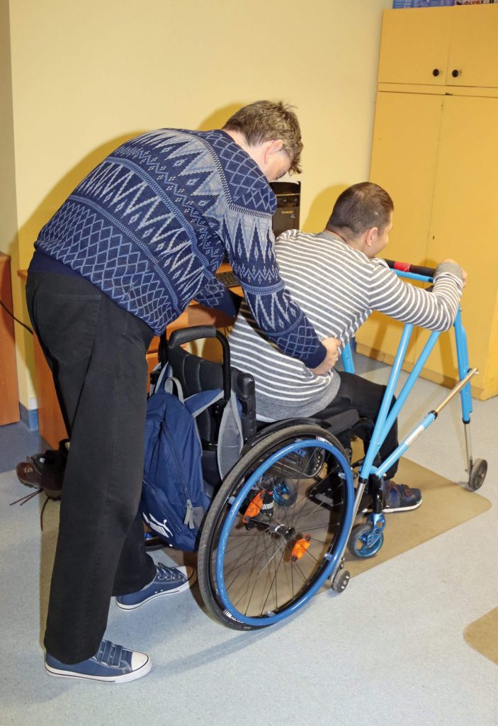 Uczestnik Warsztatu Terapii Zajęciowej w Górkach pomaga drugiemu uczestnikowi wstać z wózka inwalidzkiego