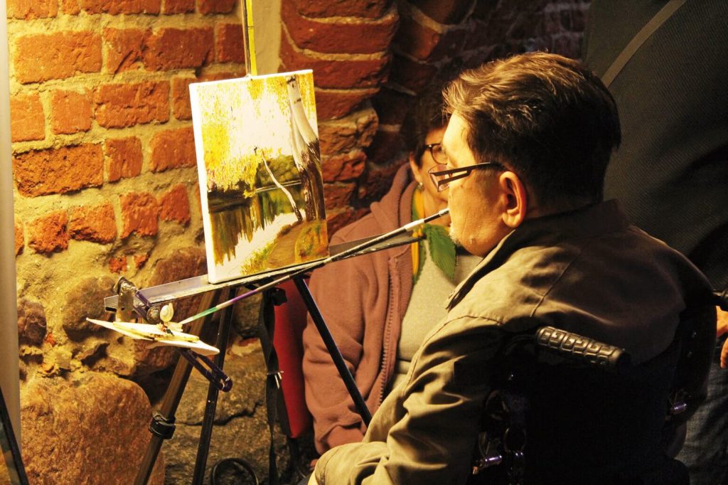 Artysta maluje obraz trzymając pędzel ustami