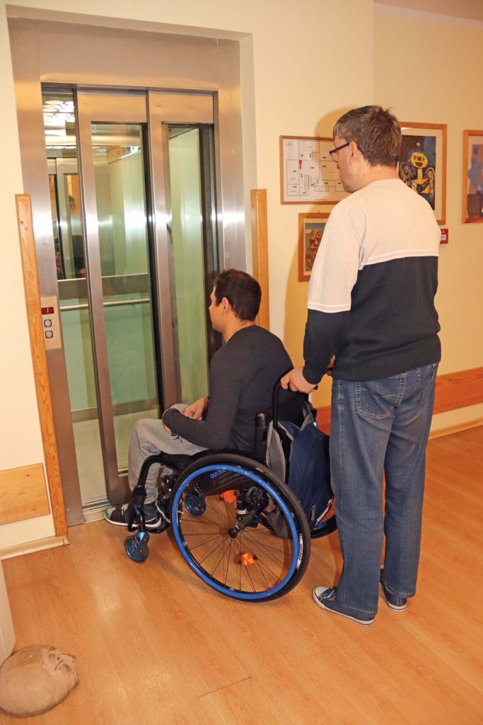Uczestnik Warsztatu Terapii Zajęciowej w Górkach prowadzi drugiego uczestnika na wózku inwalidzkim do windy