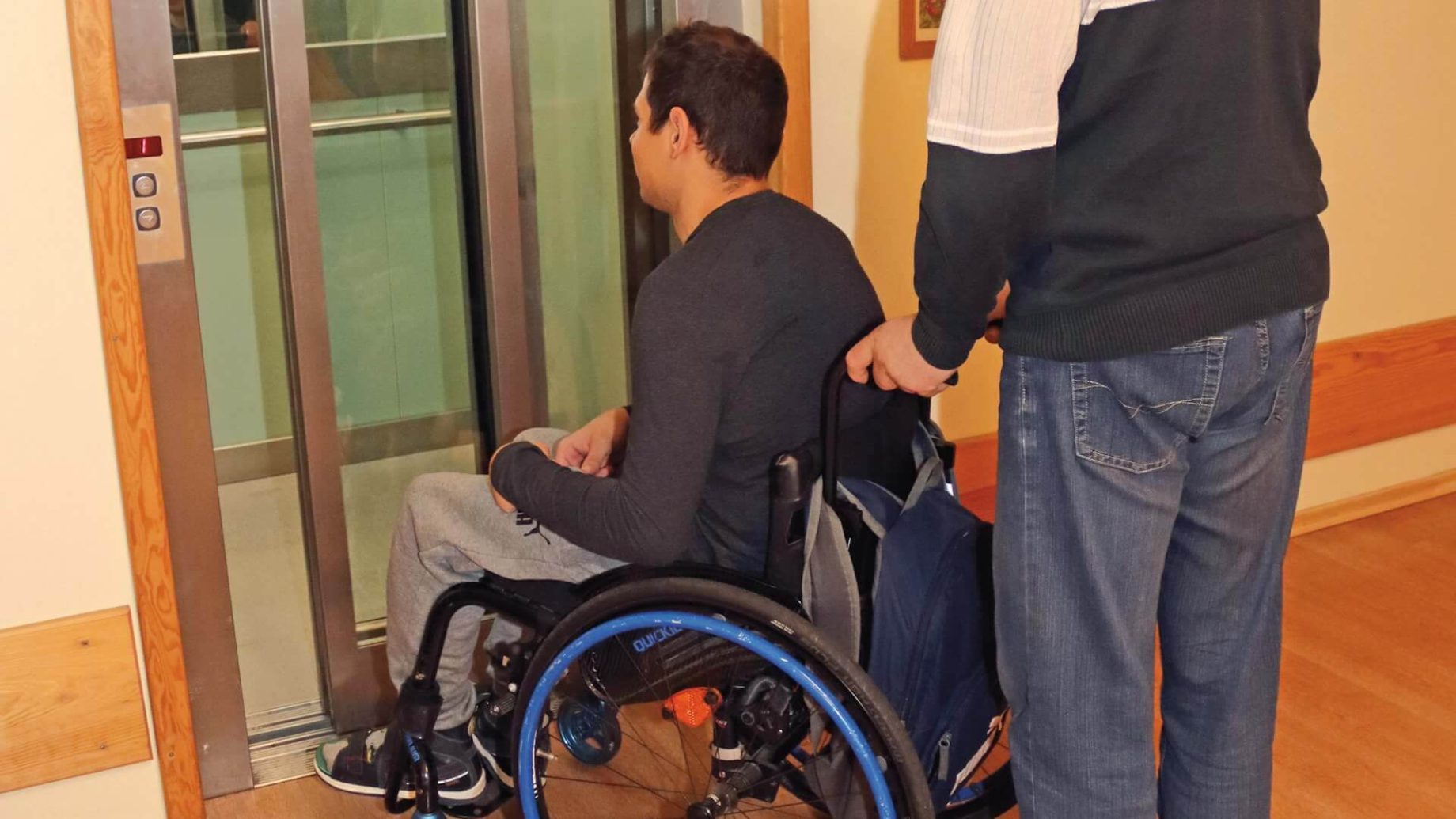 Uczestnik Warsztatu Terapii Zajęciowej w Górkach prowadzi drugiego uczestnika na wózku inwalidzkim do windy